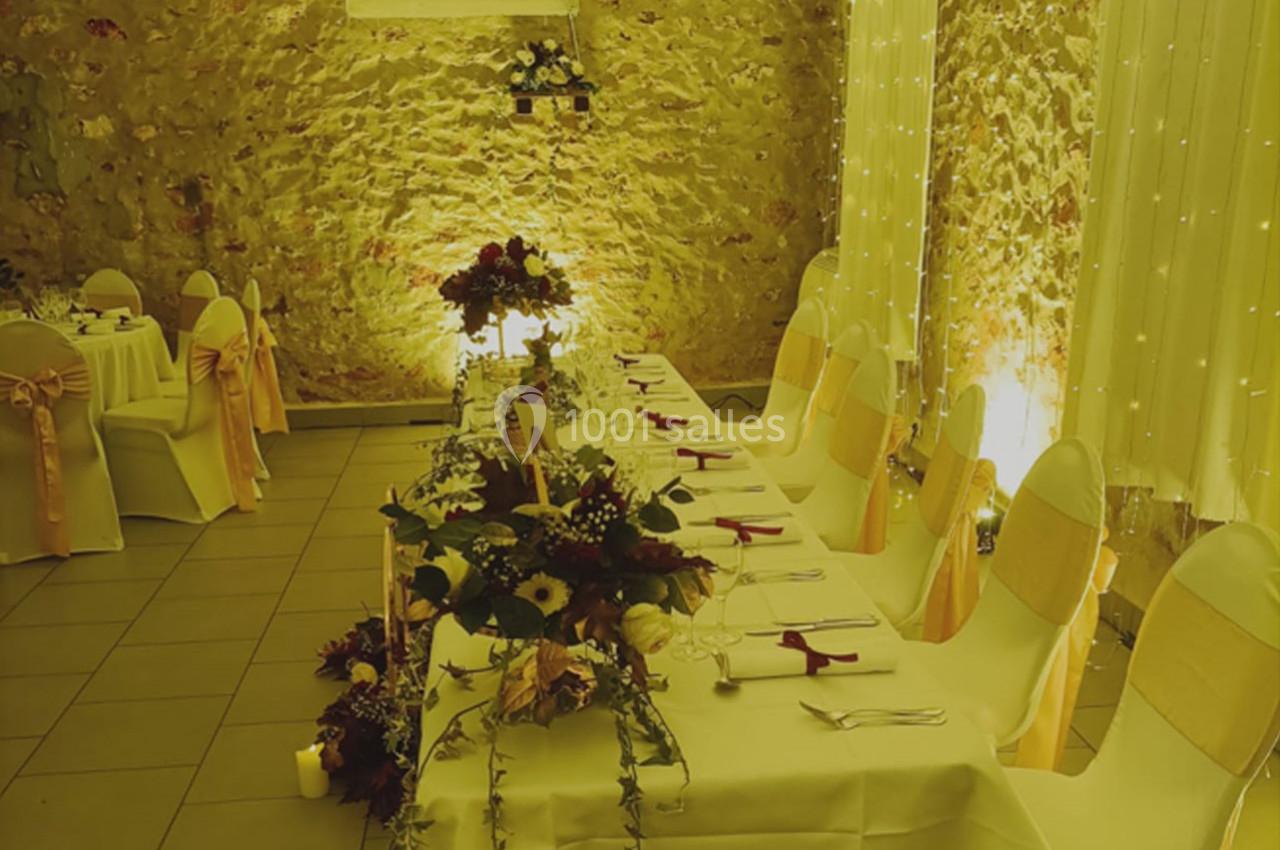 Salle de réception décorée avec des tables dressées, des chaises ornées de rubans et un éclairage chaleureux.