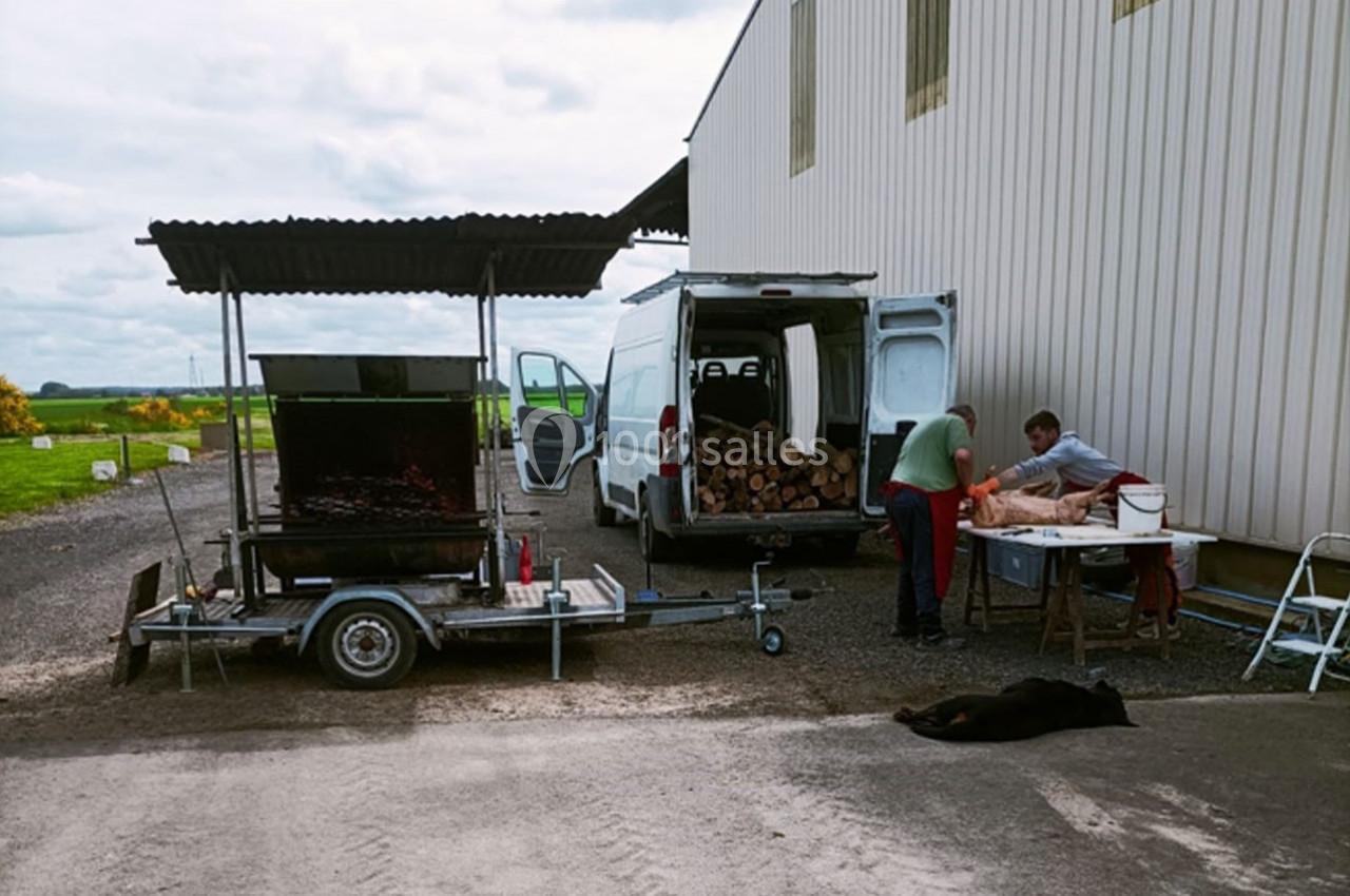 Deux personnes préparent un barbecue mobile près d'une camionnette chargée de bois, avec un chien allongé au sol.