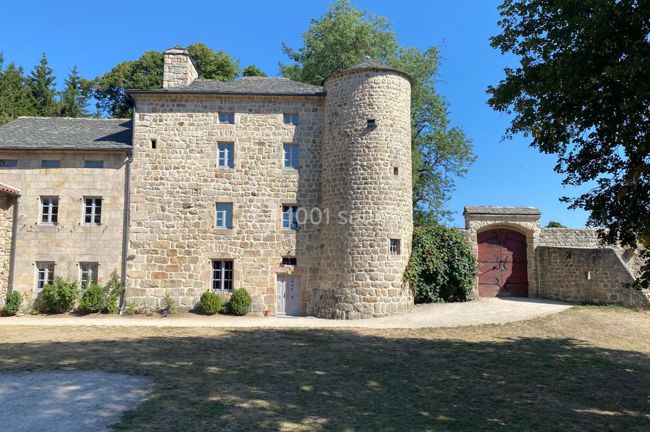 Façade en pierre d'un bâtiment historique avec une tour ronde, entouré de verdure et d'une grande porte en bois.