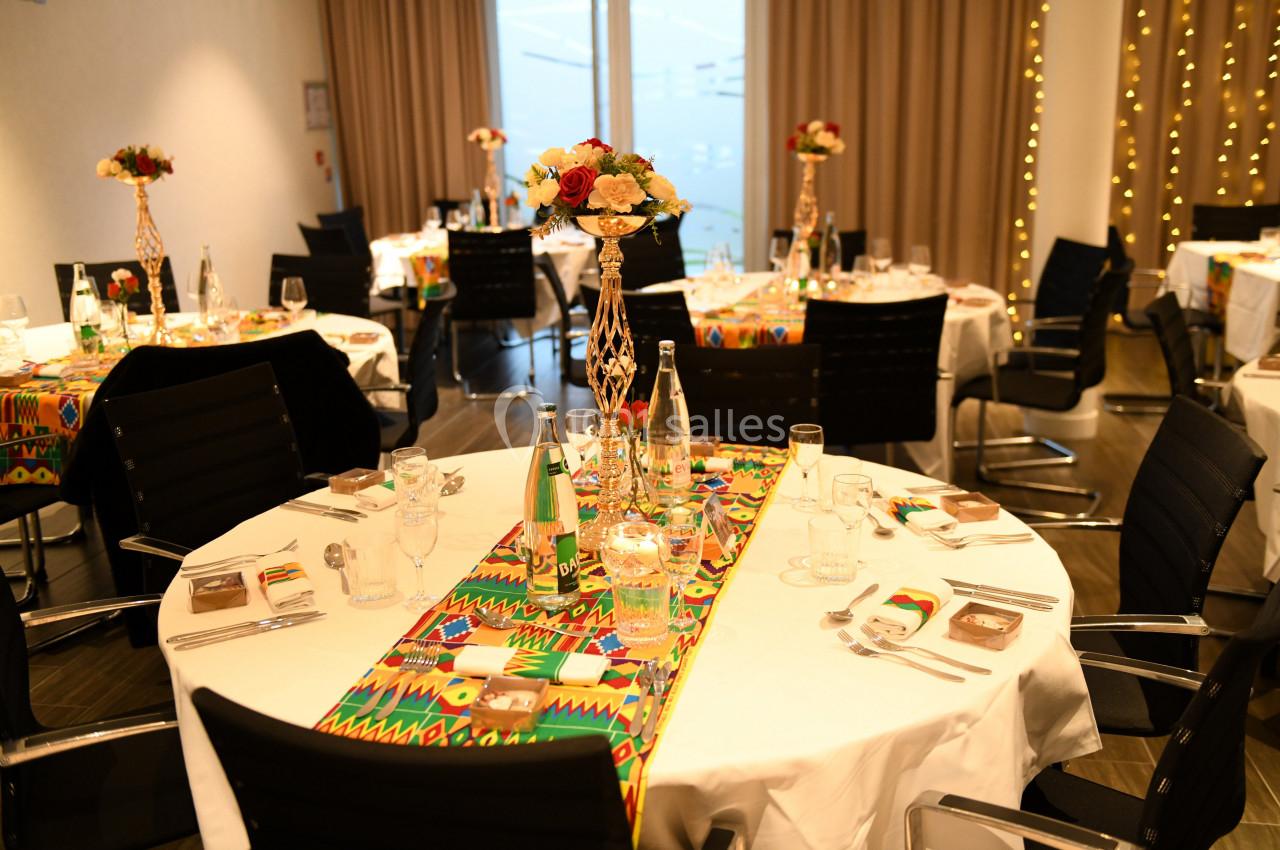 Salle décorée pour un événement avec des tables rondes, nappes blanches, chemins de table colorés et centres de table…