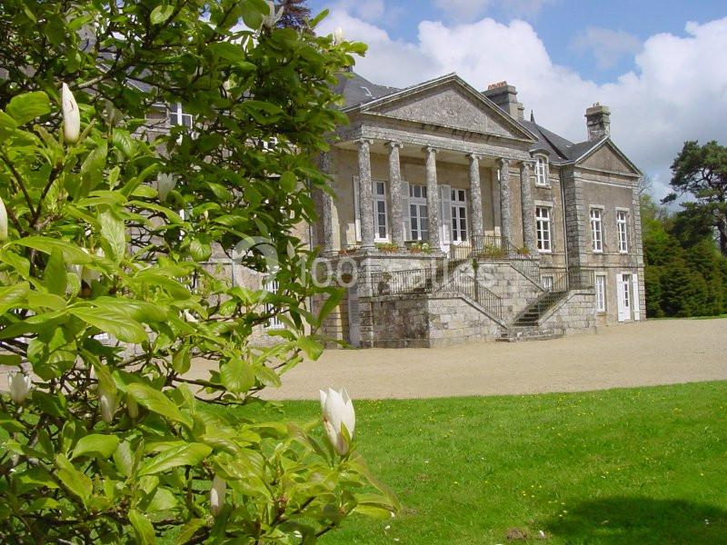Façade d'un manoir en pierre avec colonnes, entouré de verdure et d'un arbre en fleurs au premier plan.