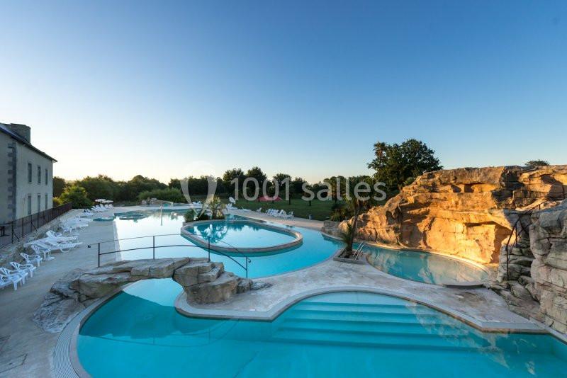 Piscine extérieure avec bassins de formes variées, entourée de rochers, transats et végétation sous un ciel dégagé.