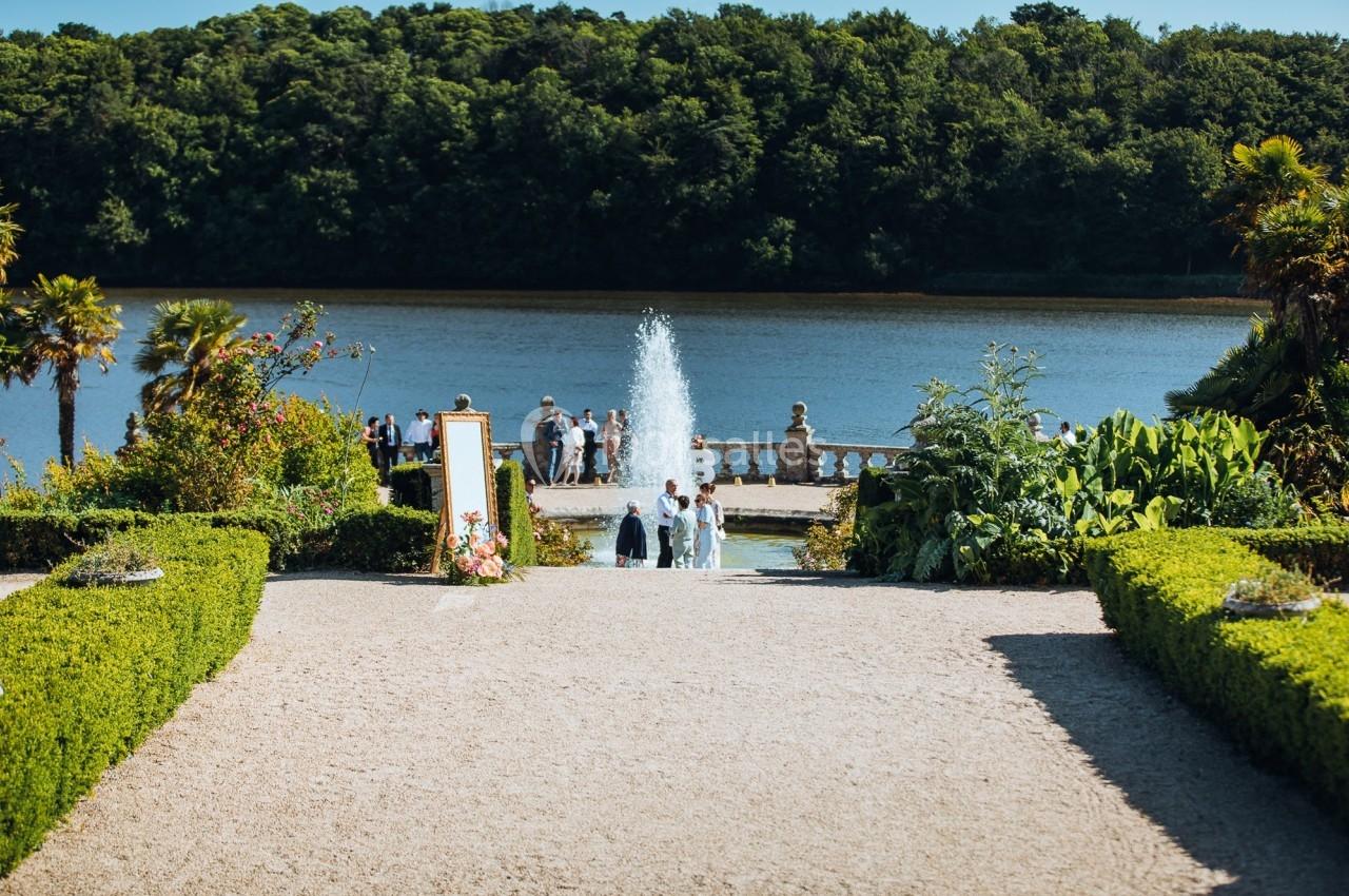 Allée bordée de verdure menant à une fontaine devant un lac, avec des invités rassemblés en arrière-plan.