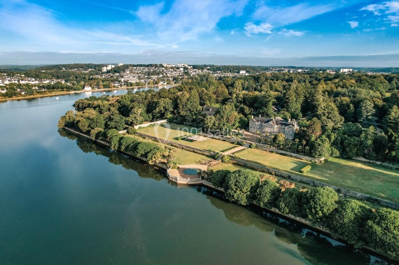 Vue aérienne d'un paysage avec une rivière, des espaces verts, des bâtiments et une ville en arrière-plan sous un ciel bleu.