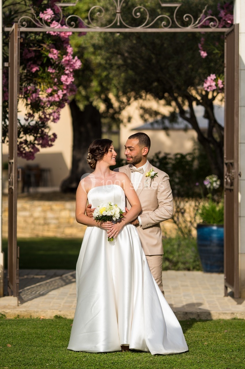 Un couple en tenue de mariage pose dans un jardin fleuri, encadré par une arche en fer forgé.