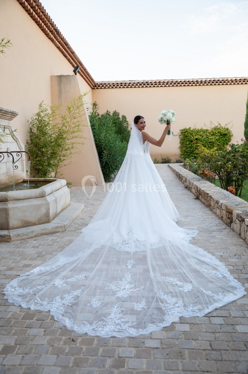 Une mariée en robe blanche avec une longue traîne tient un bouquet dans une cour pavée bordée de verdure.