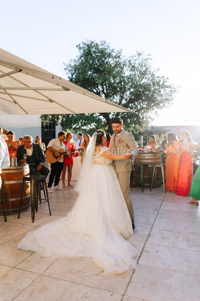 Un couple de mariés danse en extérieur lors d'une réception, entouré d'invités debout près de tables hautes.