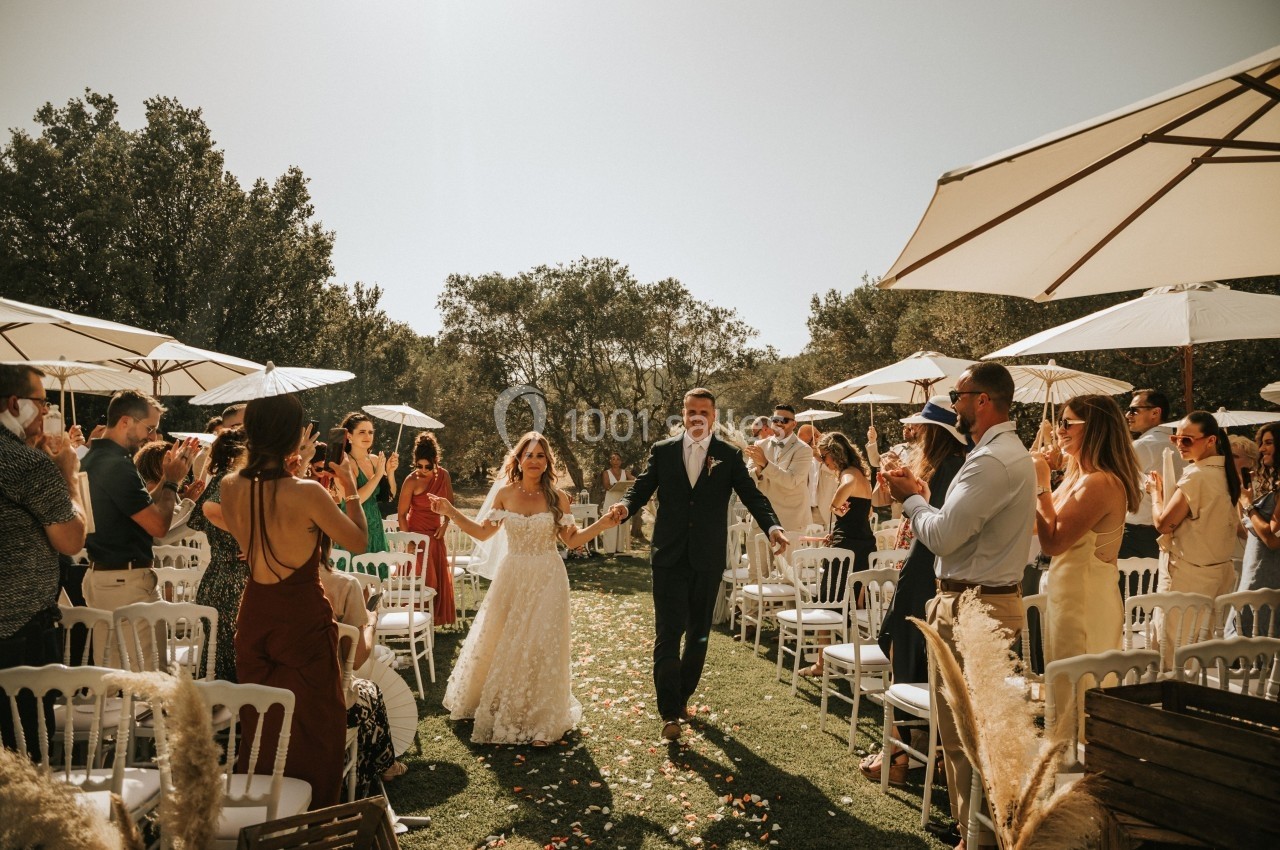 Un couple en tenue de mariage marche entre des invités applaudissant sous des parasols dans un cadre extérieur ensoleillé.