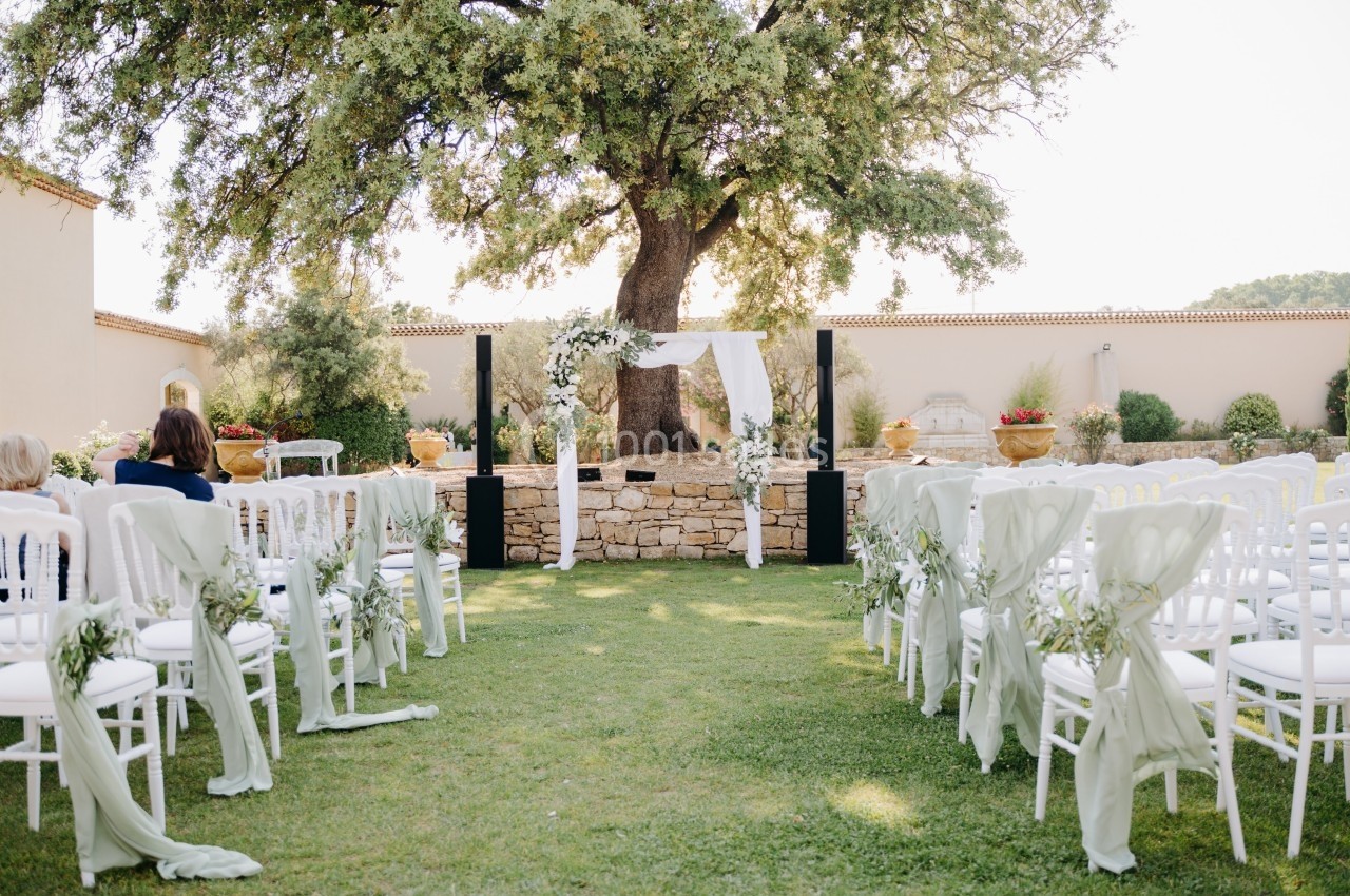 Chaises blanches décorées alignées face à une arche de mariage sous un grand arbre dans un jardin ensoleillé.