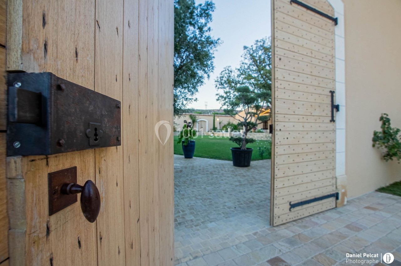 Porte en bois ouverte donnant sur une cour pavée avec des arbres et des plantes en pot.