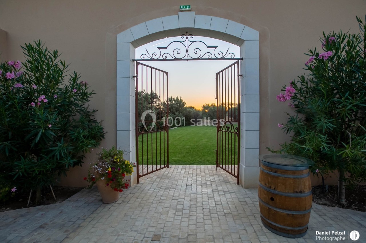 Portail en fer forgé ouvert sur un jardin verdoyant au coucher du soleil, entouré de plantes et d'un tonneau en bois.
