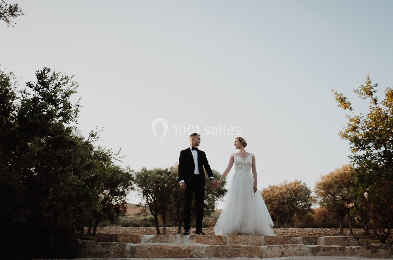 Un couple en tenue de mariage se tient la main sur des marches en pierre, entouré d'arbres sous un ciel clair.