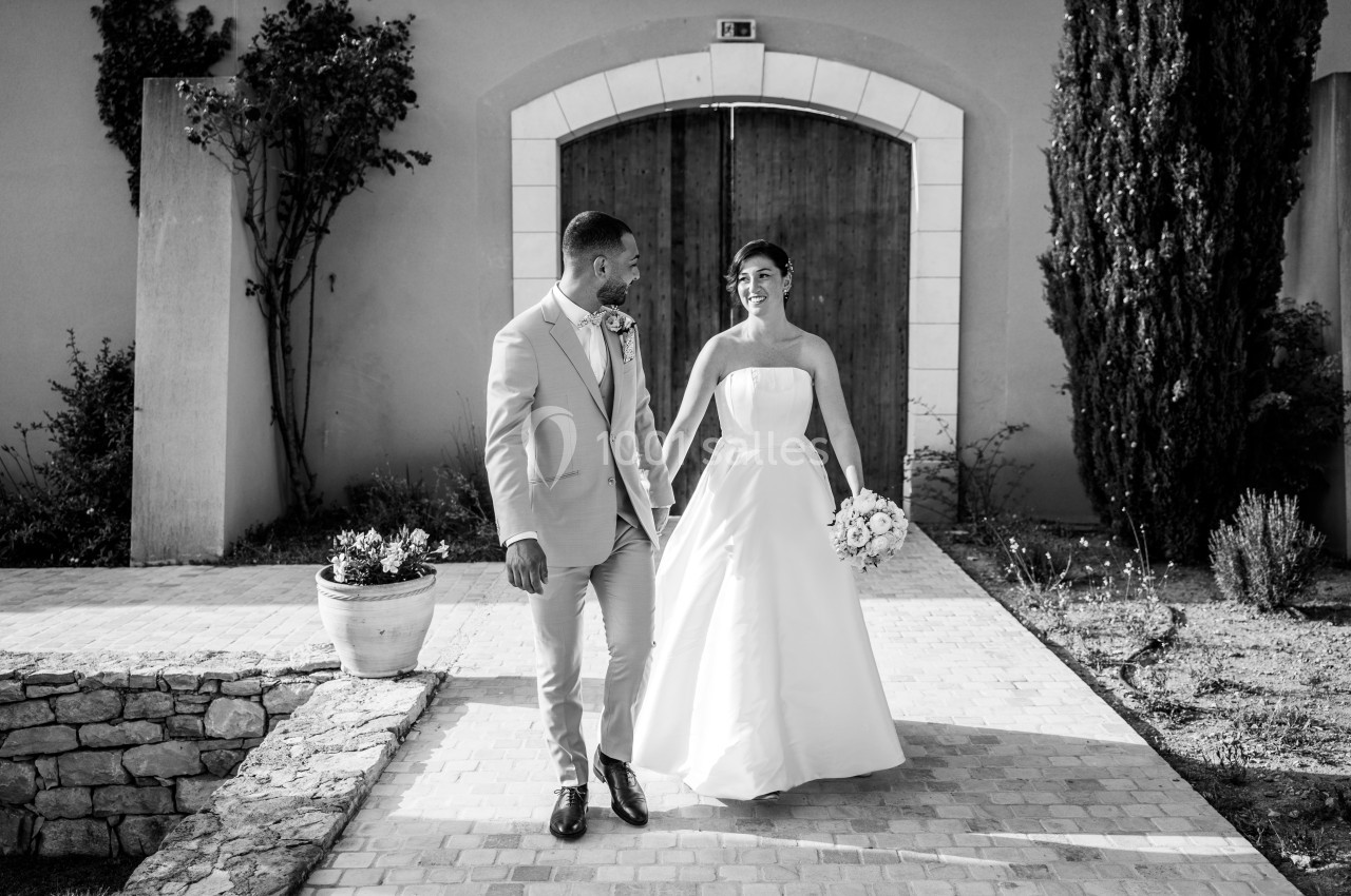Un couple en tenue de mariage marche devant une porte en bois dans une cour pavée, souriant l'un à l'autre.