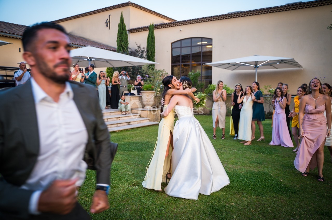 Deux femmes en robe de soirée s'embrassent dans un jardin lors d'une fête, entourées d'invités souriants.