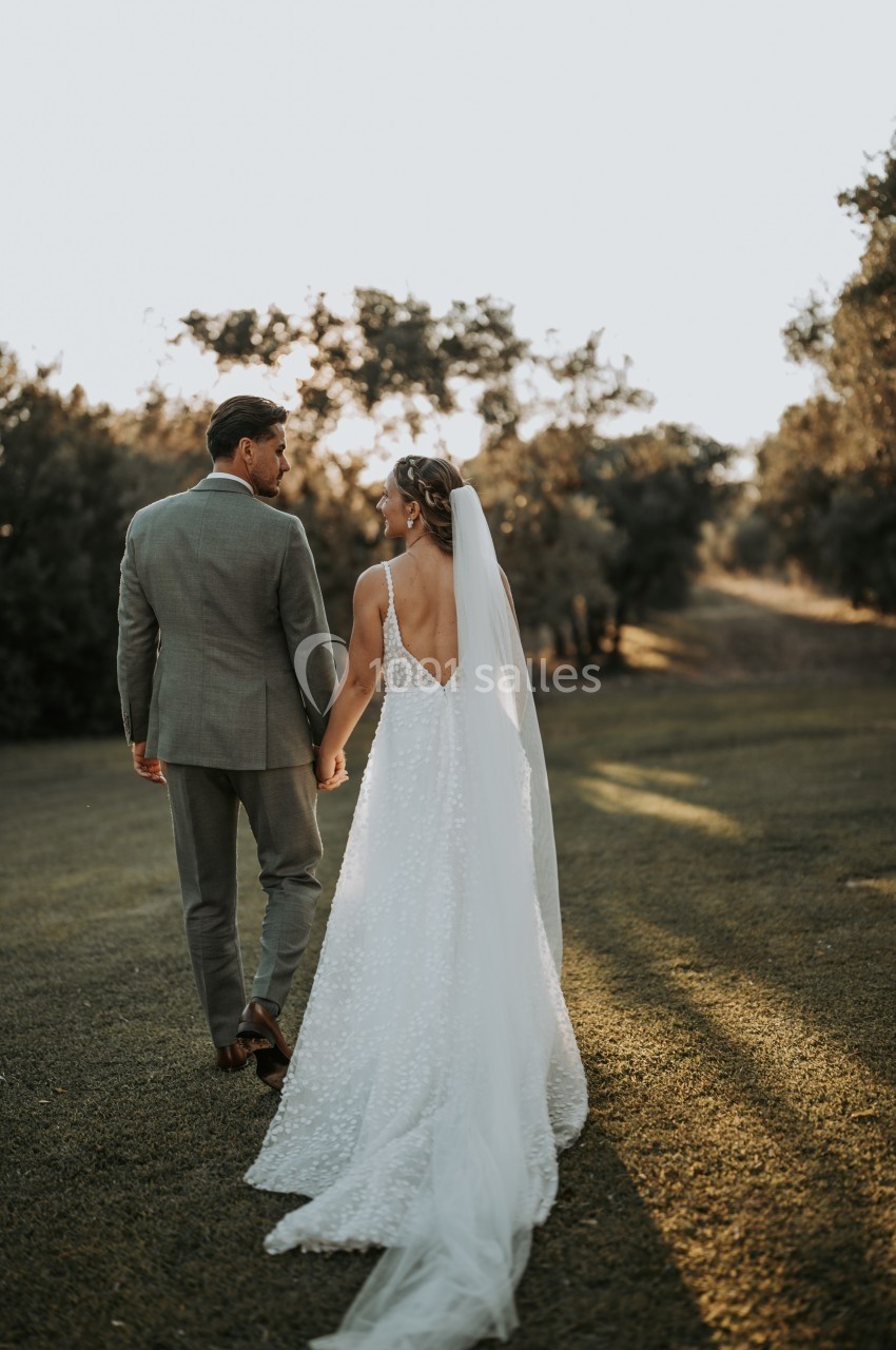 Un couple en tenue de mariage marche main dans la main sur une pelouse au coucher du soleil.