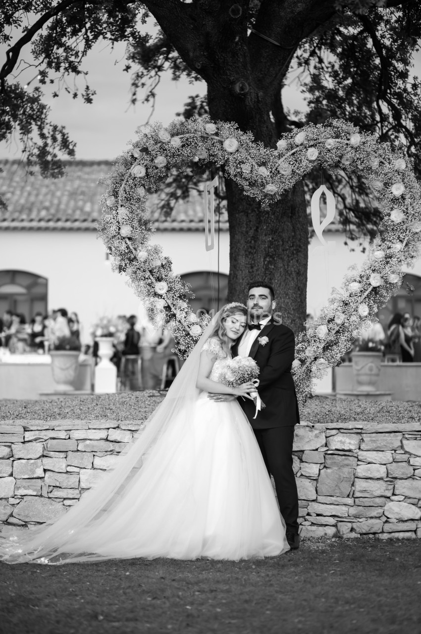Un couple de mariés pose sous un arbre décoré d'un grand cœur floral, avec des invités en arrière-plan flou.