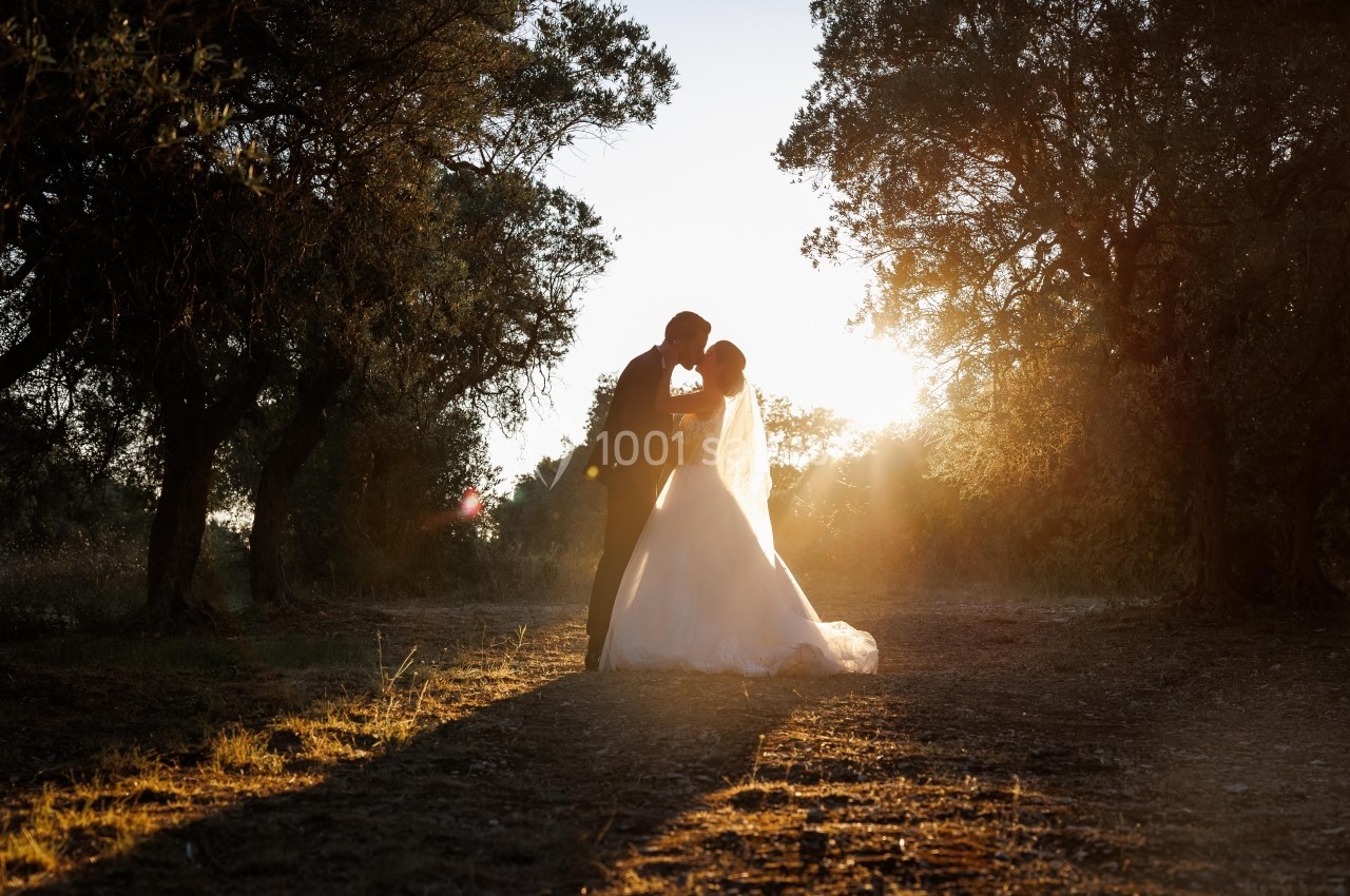 Un couple en tenue de mariage s'embrasse dans un chemin boisé au coucher du soleil.