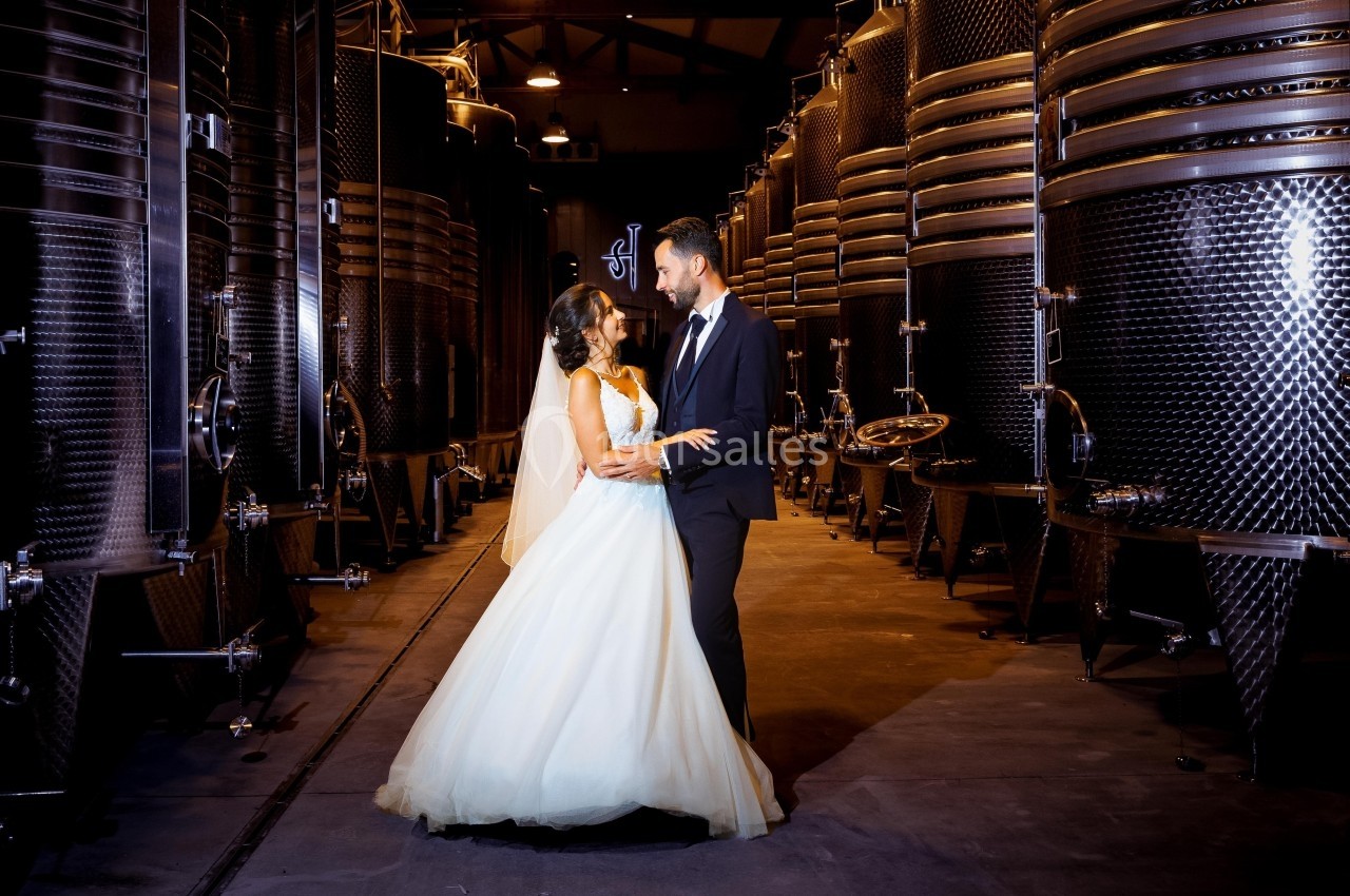 Un couple en tenue de mariage pose dans une cave entourée de grandes cuves métalliques.