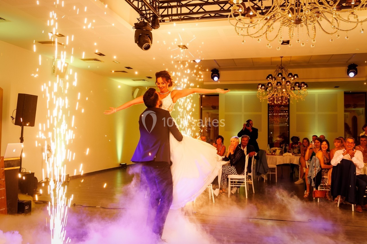 Un couple danse au centre d'une salle illuminée, entouré d'effets pyrotechniques et de fumée, sous le regard des invités.