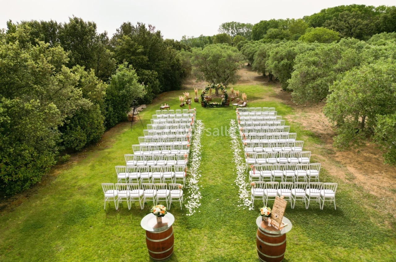 Chaises blanches alignées en extérieur pour une cérémonie, avec une arche fleurie au fond et des arbres environnants.