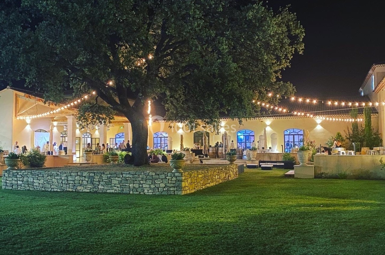 Grande maison éclairée la nuit avec des guirlandes lumineuses, un grand arbre et une pelouse au premier plan.