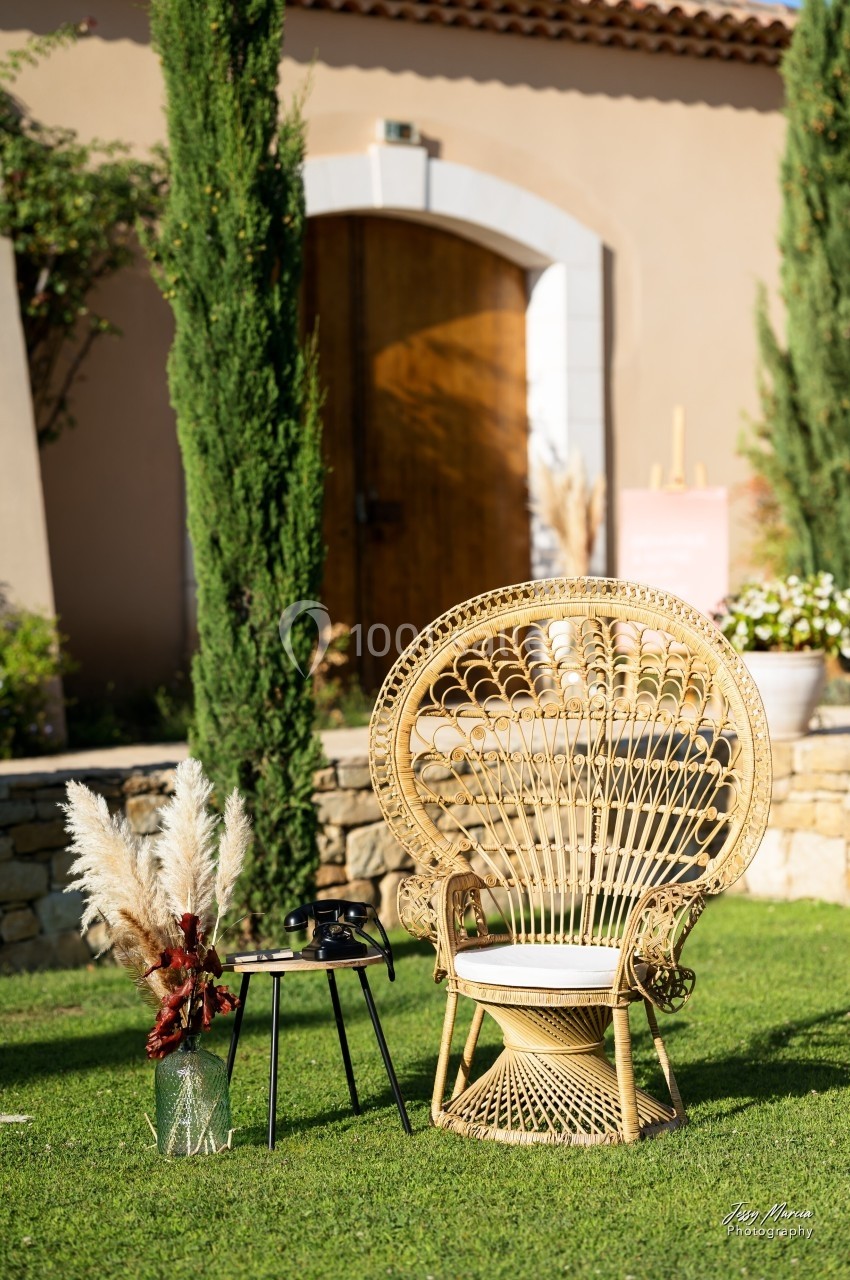 Fauteuil en rotin avec table basse et vase de pampas, installé sur une pelouse devant une maison en pierre.