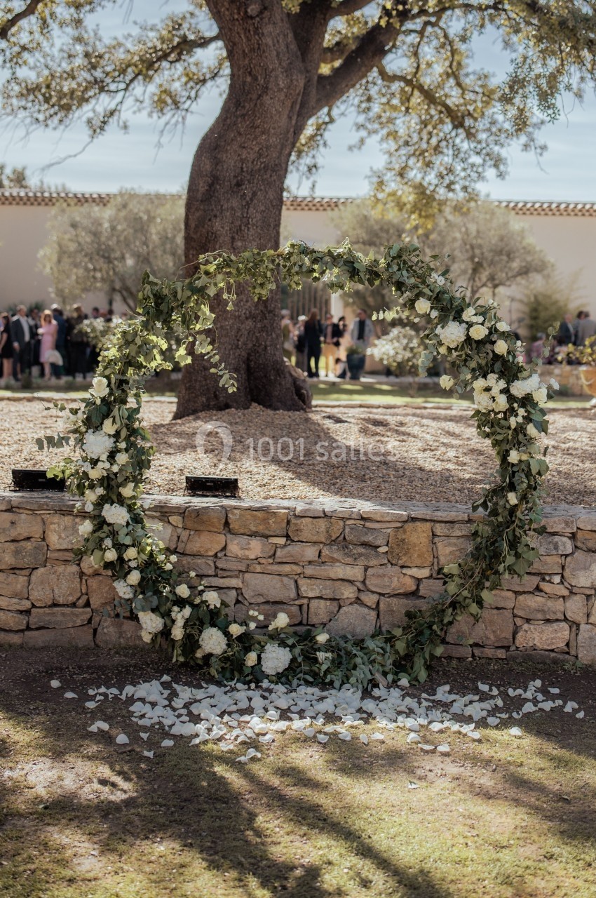 Cercle floral décoratif en feuillage et fleurs blanches, posé devant un muret en pierre dans un jardin ensoleillé.