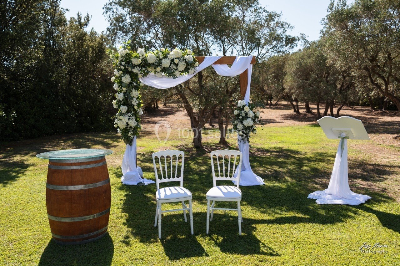 Arche fleurie avec deux chaises blanches, un tonneau en bois et un pupitre dans un jardin ensoleillé.