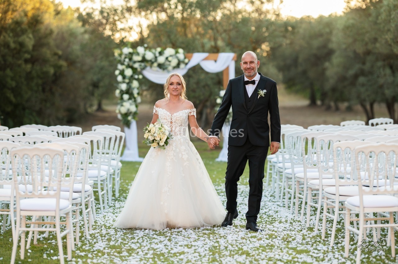 Un couple en tenue de mariage marche main dans la main dans une allée bordée de chaises blanches, décorée de pétales.