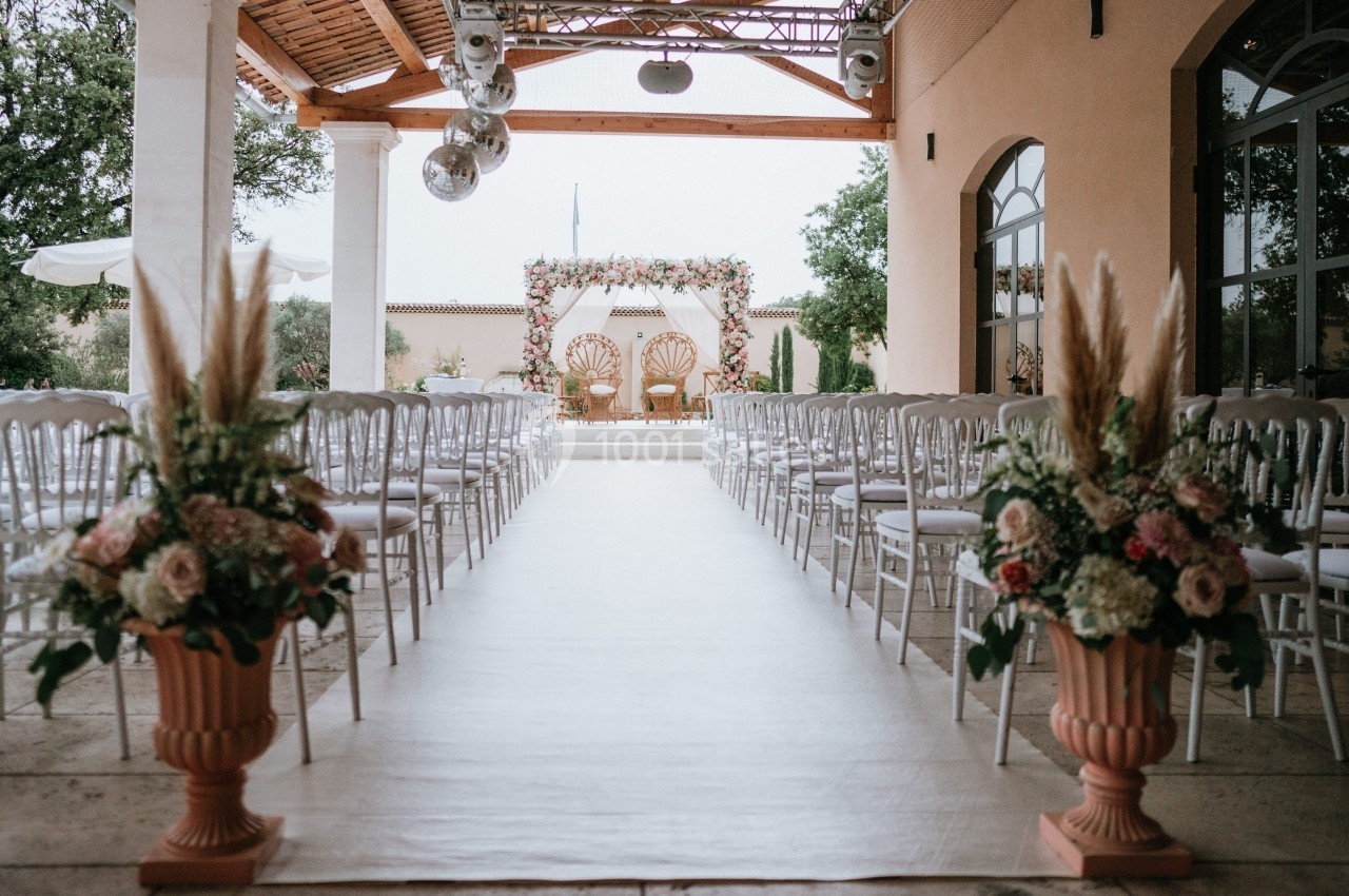 Allée centrale décorée de fleurs menant à une arche ornée pour une cérémonie en extérieur sous une pergola.