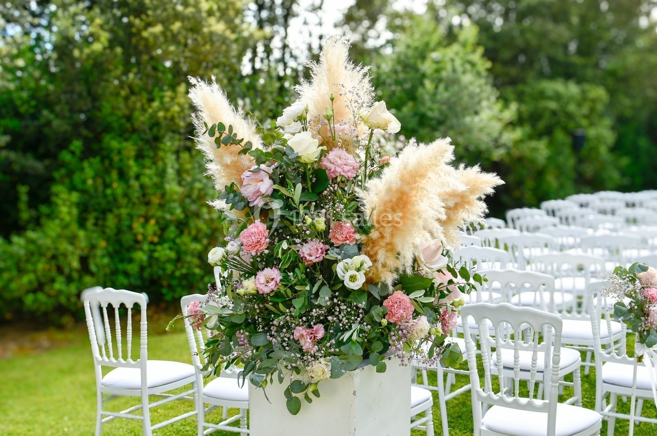 Arrangement floral avec roses, pampas et feuillage, placé devant des chaises blanches alignées dans un jardin.