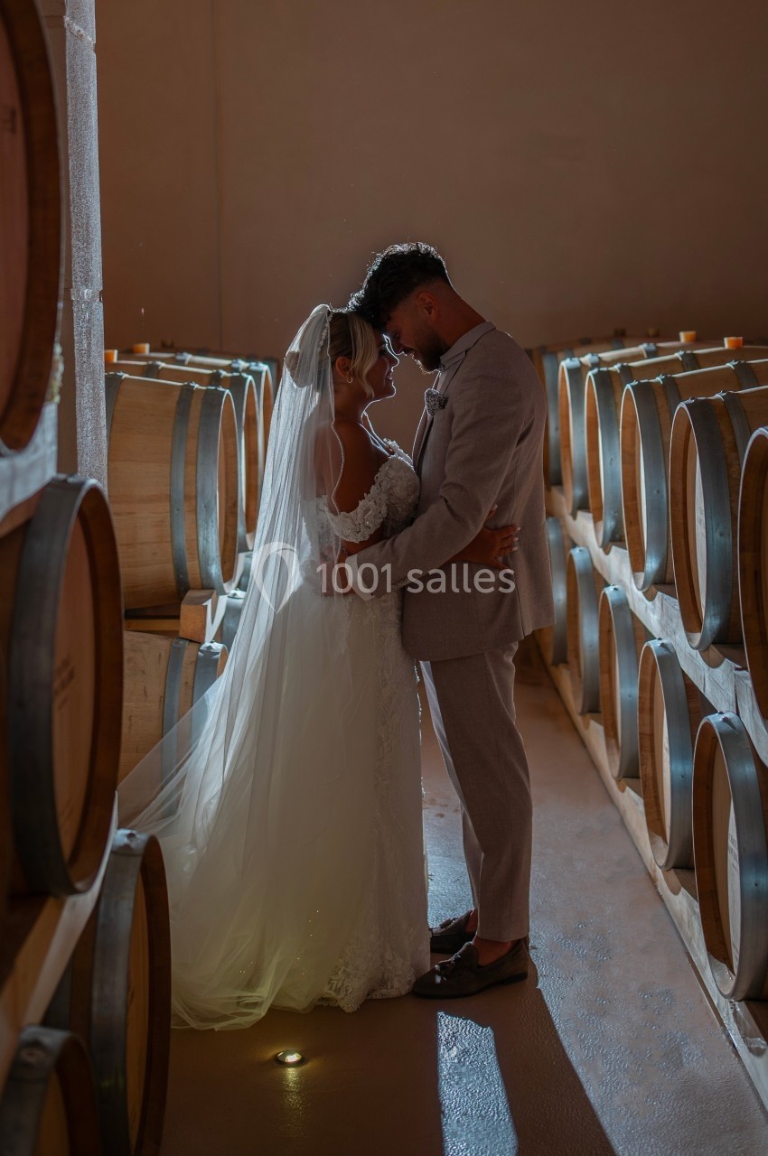 Un couple en tenue de mariage se tient enlacé dans une cave à vin éclairée par une lumière douce.
