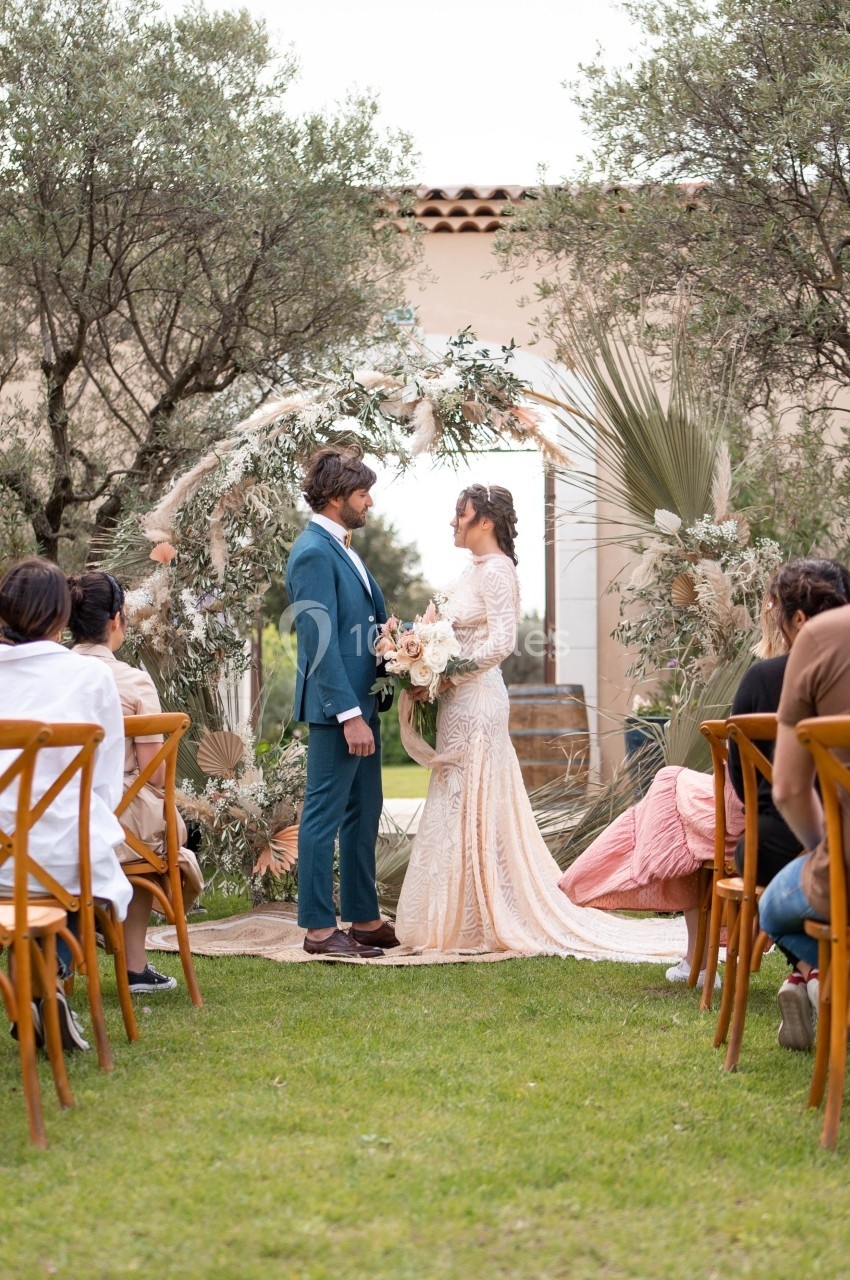 Un couple se tient devant une arche florale lors d'une cérémonie de mariage en extérieur, entouré d'invités assis.
