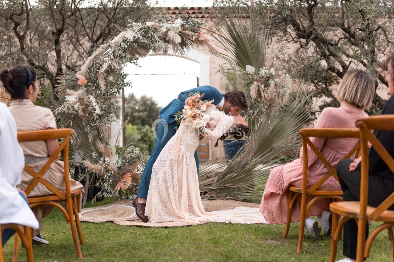 Un couple échange un baiser sous une arche décorée de fleurs séchées lors d'une cérémonie en plein air.