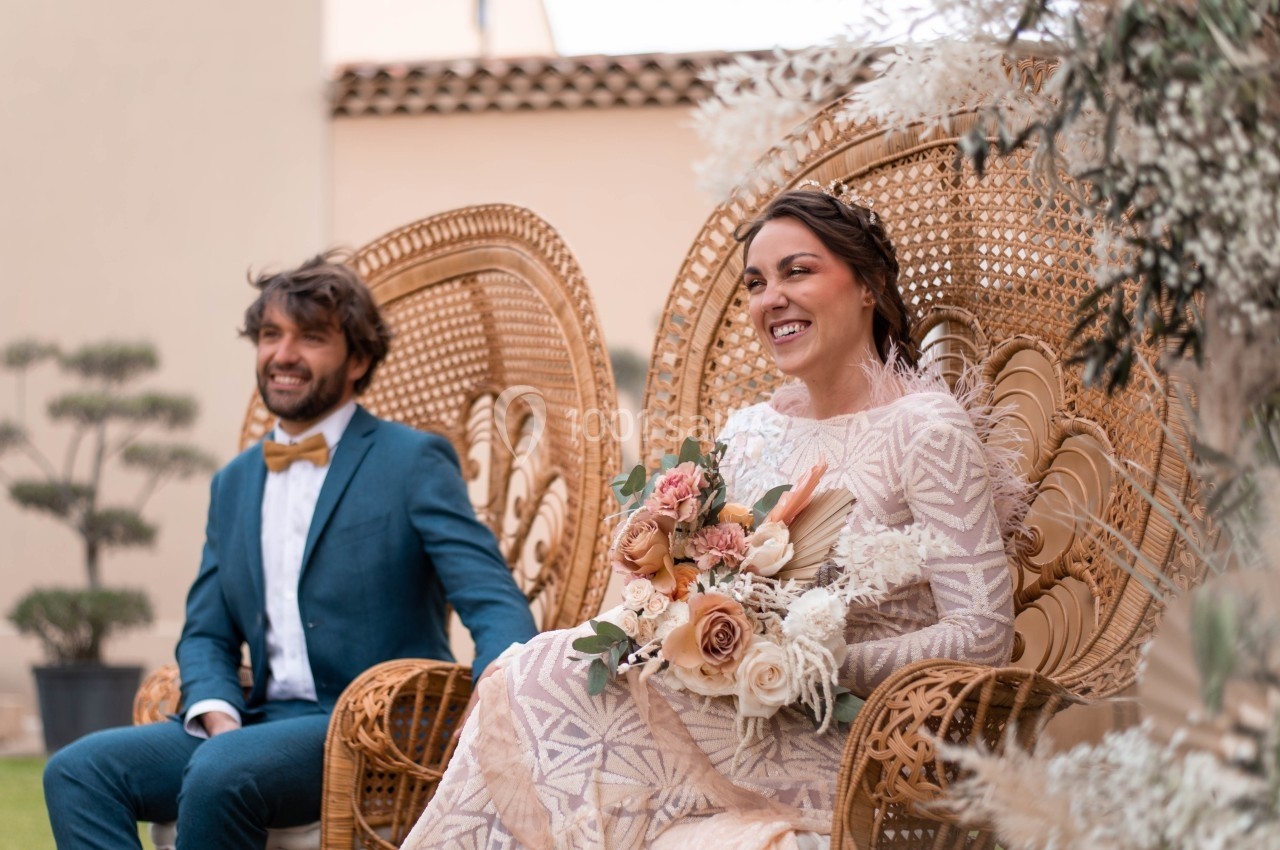 Un couple assis sur des fauteuils en rotin, la mariée tenant un bouquet de fleurs, dans un cadre extérieur lumineux.