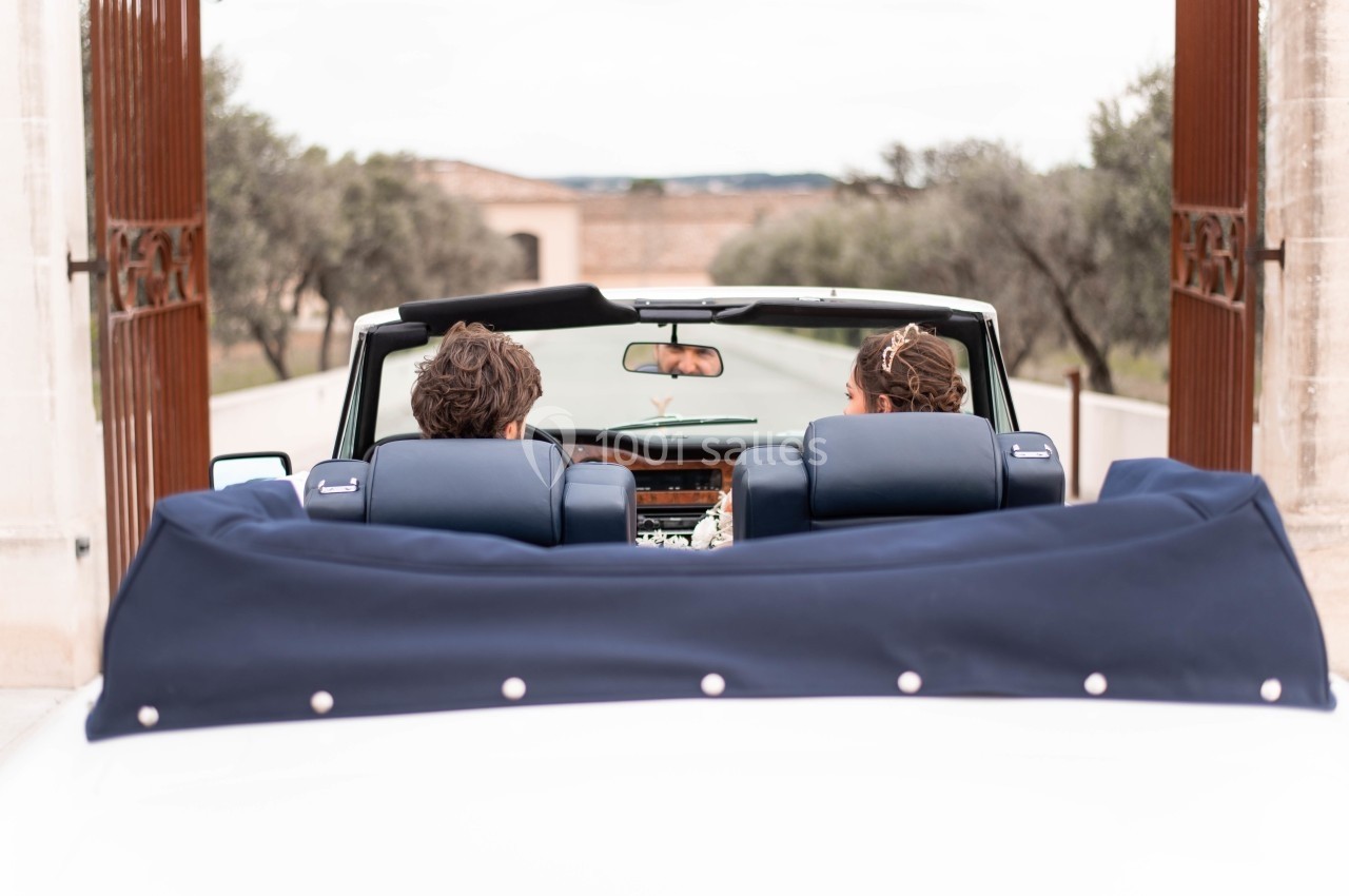 Deux personnes assises dans une voiture décapotable blanche roulant sur une route bordée d'oliviers.