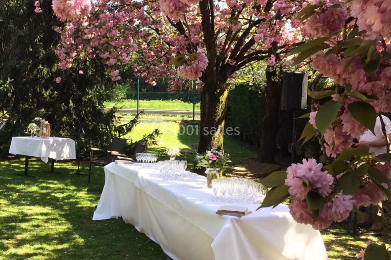 Table dressée sous des cerisiers en fleurs dans un jardin, avec nappes blanches et verres disposés.