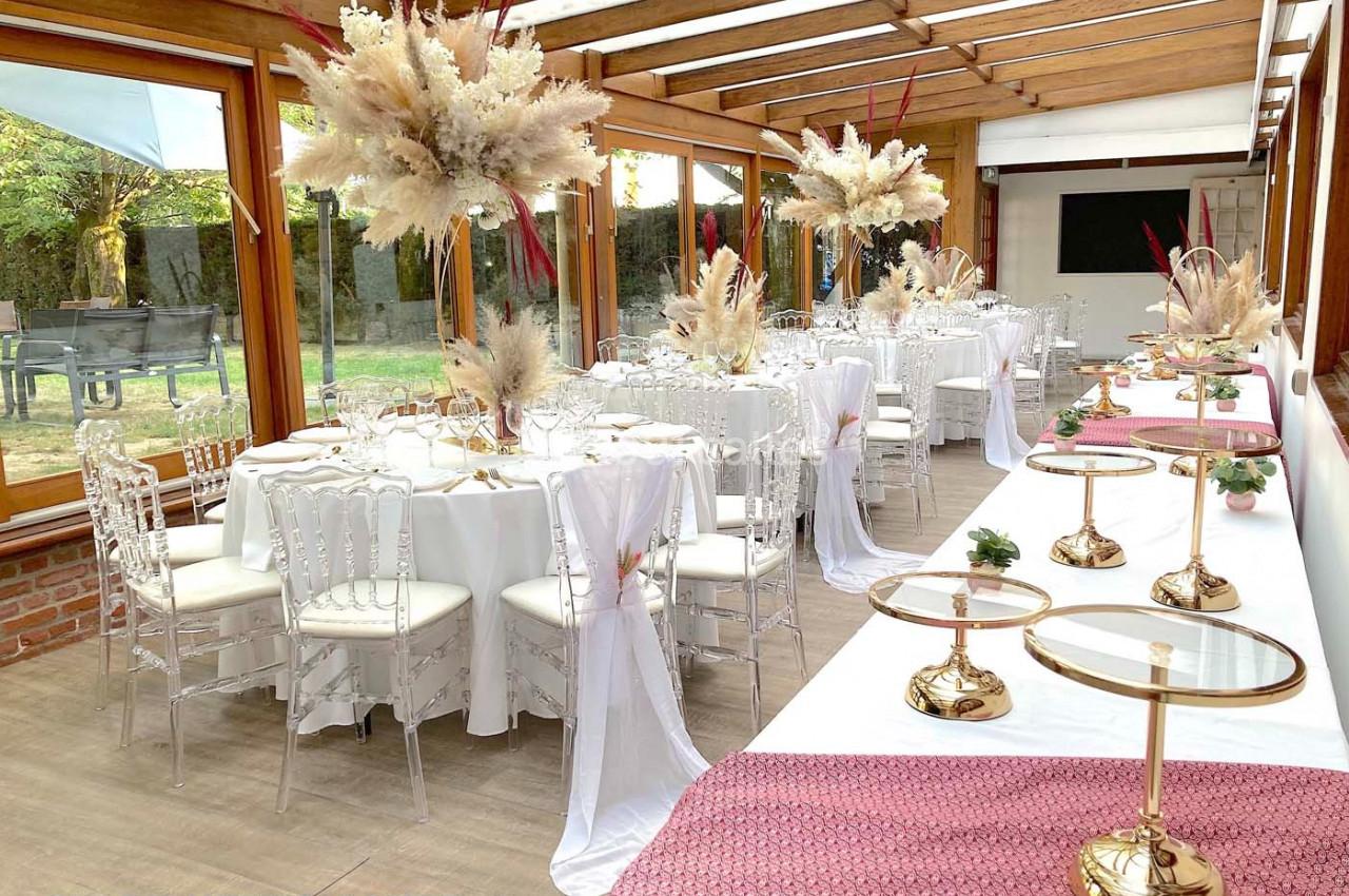 Salle décorée pour un événement avec tables dressées, chaises transparentes et compositions florales sous une véranda…