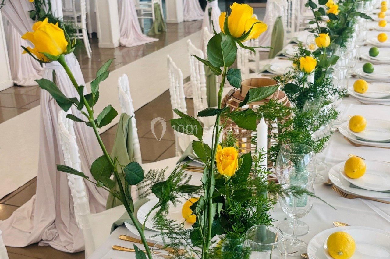 Table décorée avec des roses jaunes, des citrons et des éléments verts dans une salle lumineuse aux rideaux blancs.