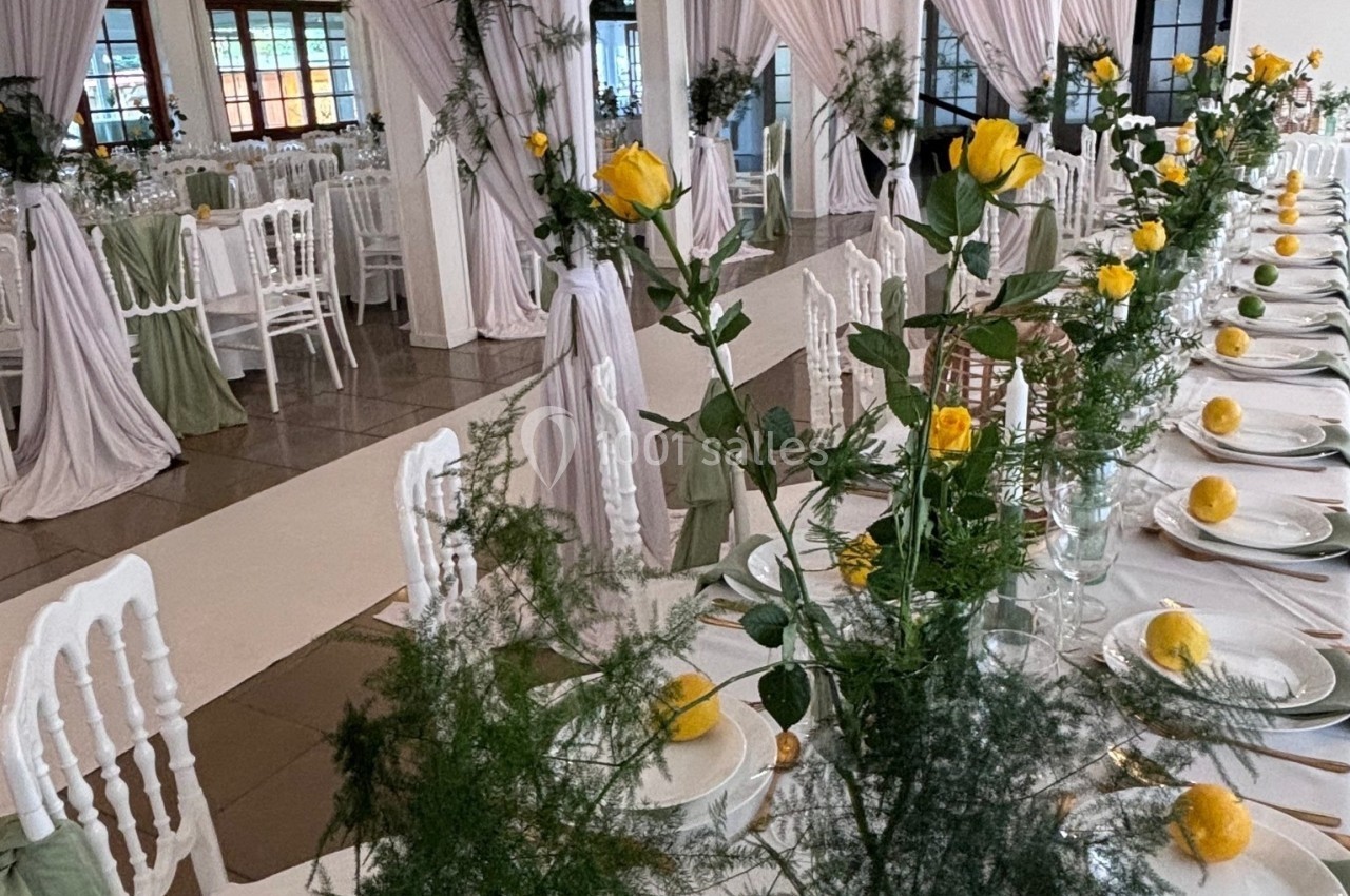 Salle de réception décorée avec des tables alignées, nappes blanches, roses jaunes et feuillage vert.