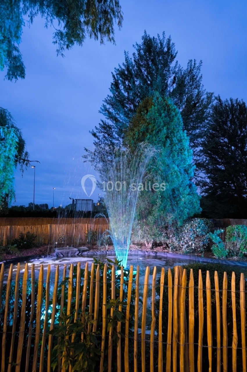 Fontaine éclairée la nuit dans un jardin entouré de palissades en bois, avec des arbres en arrière-plan.