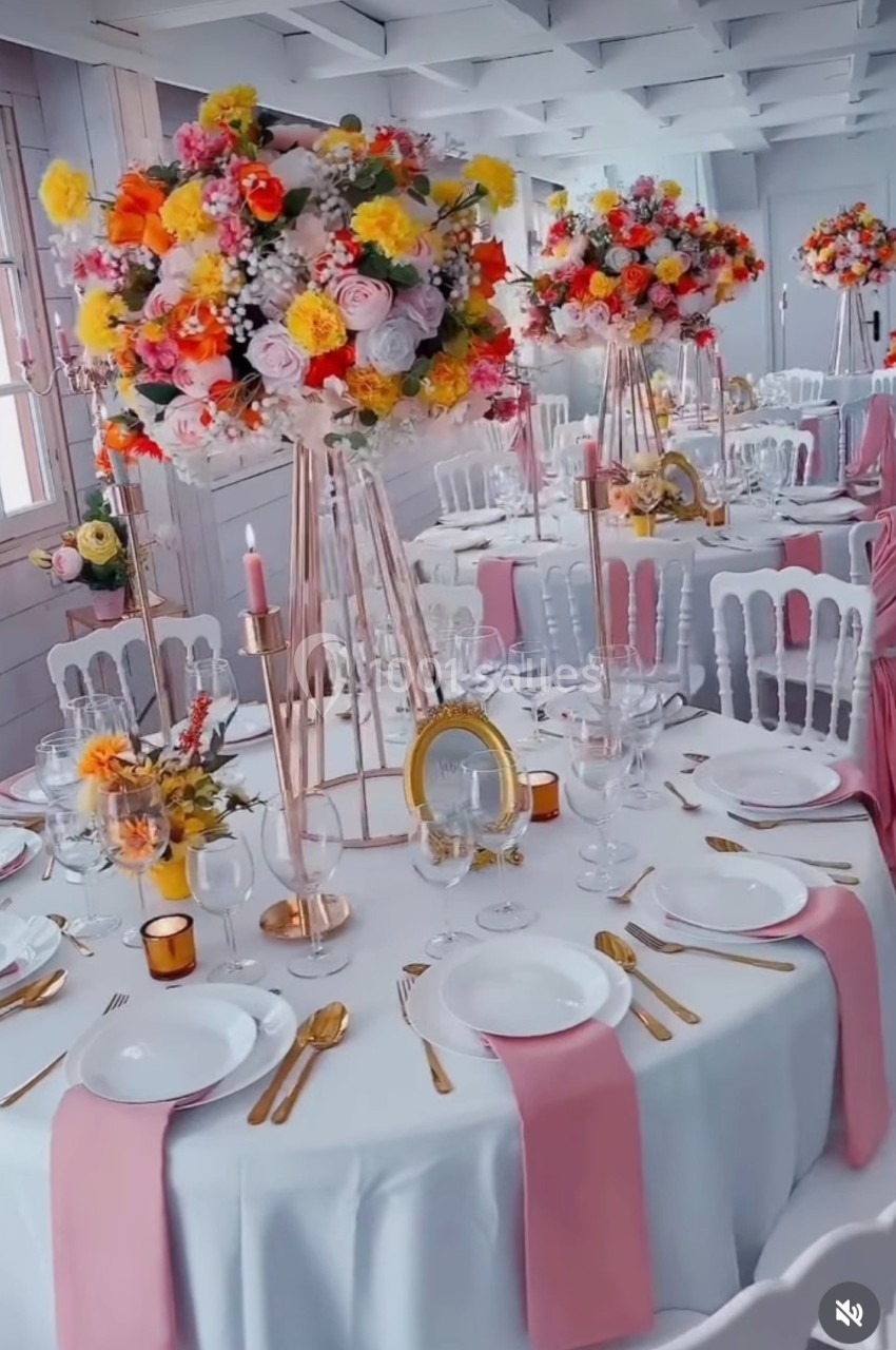 Salle de réception décorée avec des tables rondes, nappes blanches, serviettes roses et centres de table fleuris colorés.
