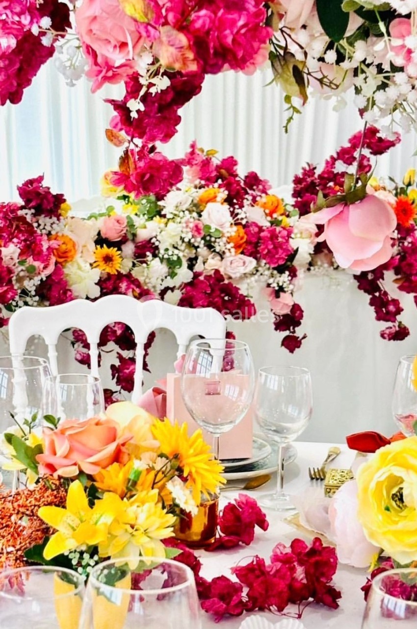 Table décorée avec des fleurs colorées, des verres et des assiettes, entourée d'une abondance de compositions florales.