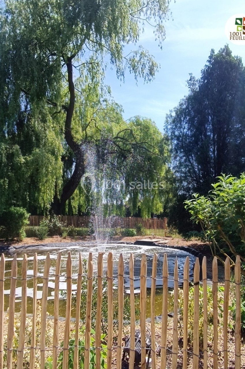 Étang entouré de végétation avec une fontaine centrale, clôturé par une barrière en bois sous un ciel dégagé.