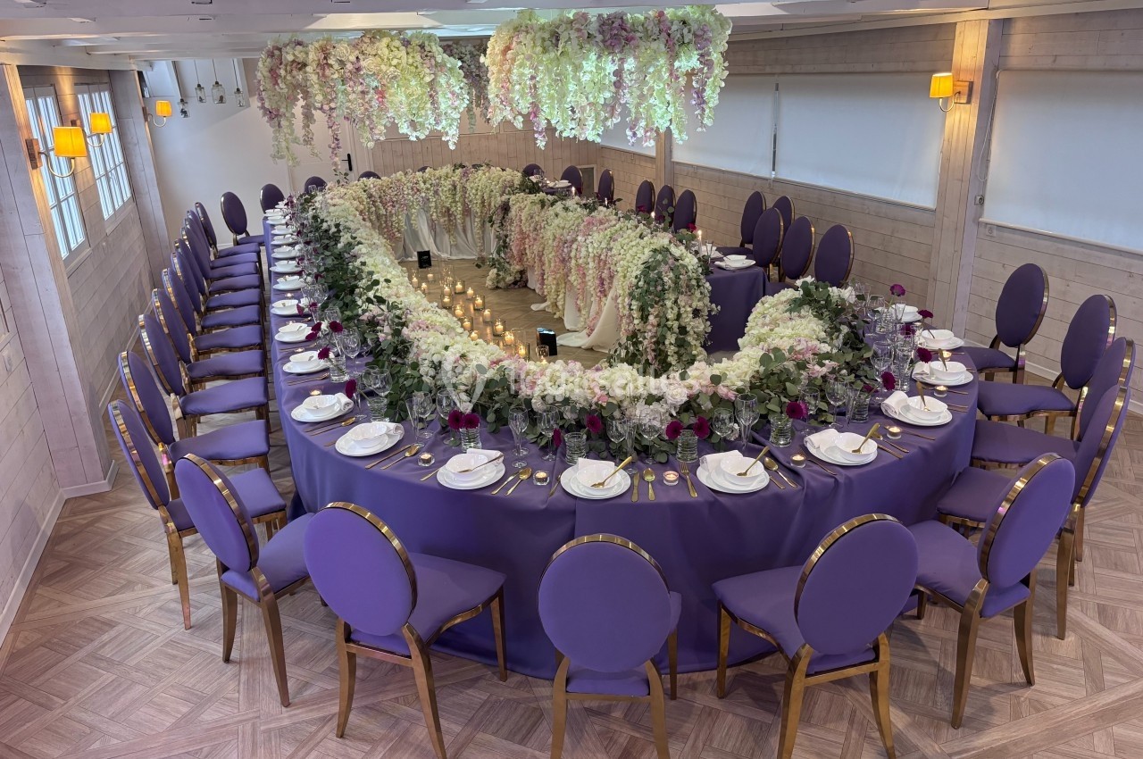 Salle de réception avec une grande table ovale décorée de fleurs blanches et violettes, nappes violettes et chaises…