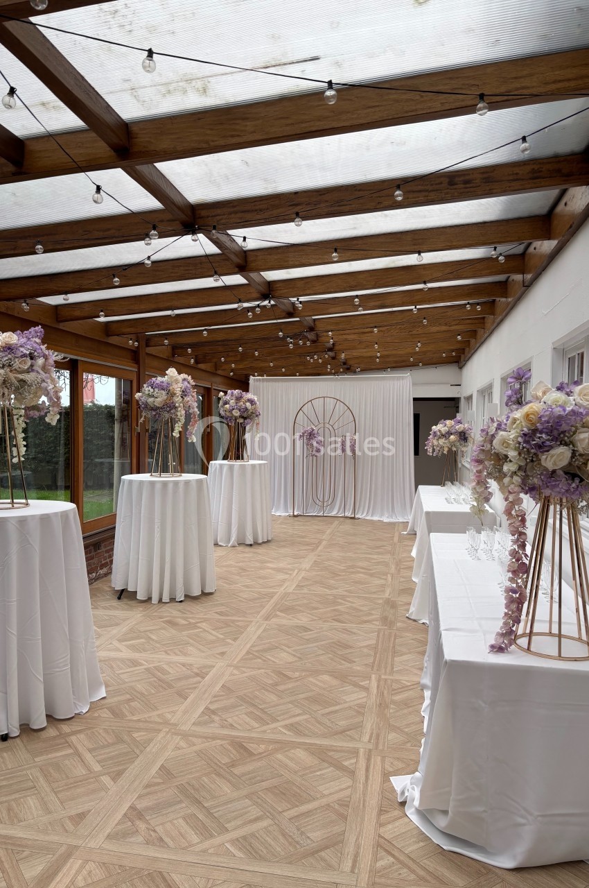 Salle décorée pour un événement, avec tables hautes drapées de blanc et ornées de bouquets pastel sous une verrière.