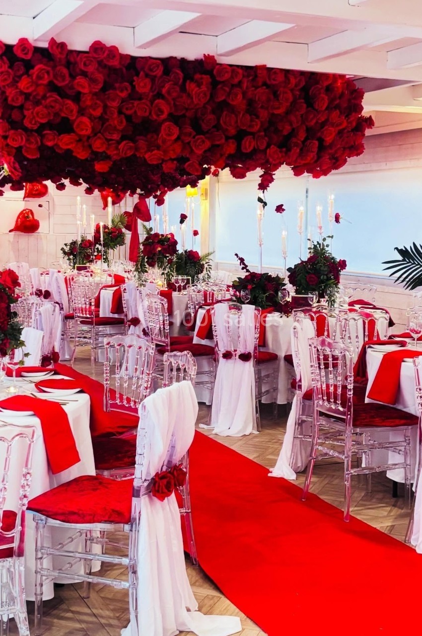 Salle de réception décorée avec des roses rouges, nappes blanches, chaises transparentes et tapis rouge au centre.