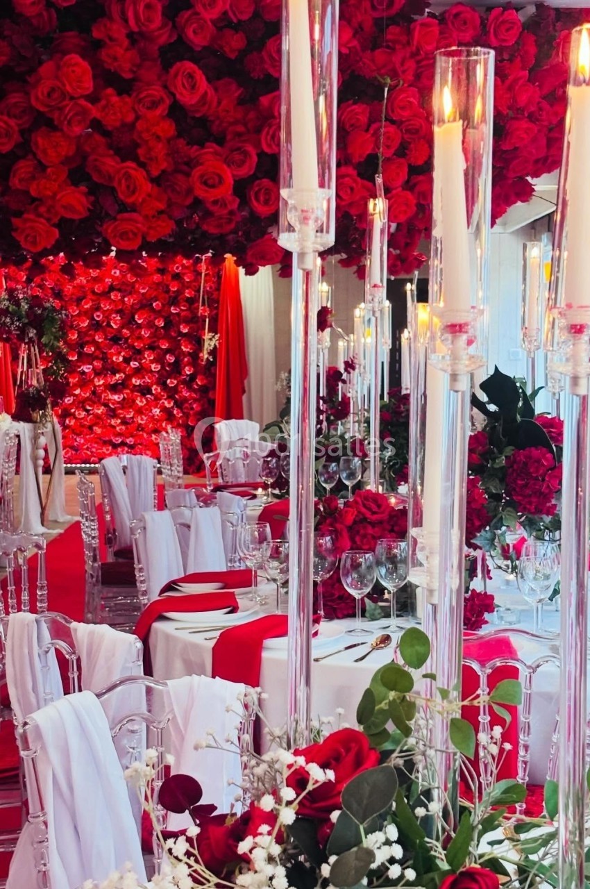 Salle de réception décorée de roses rouges, chandeliers en verre et nappes blanches avec accents rouges.