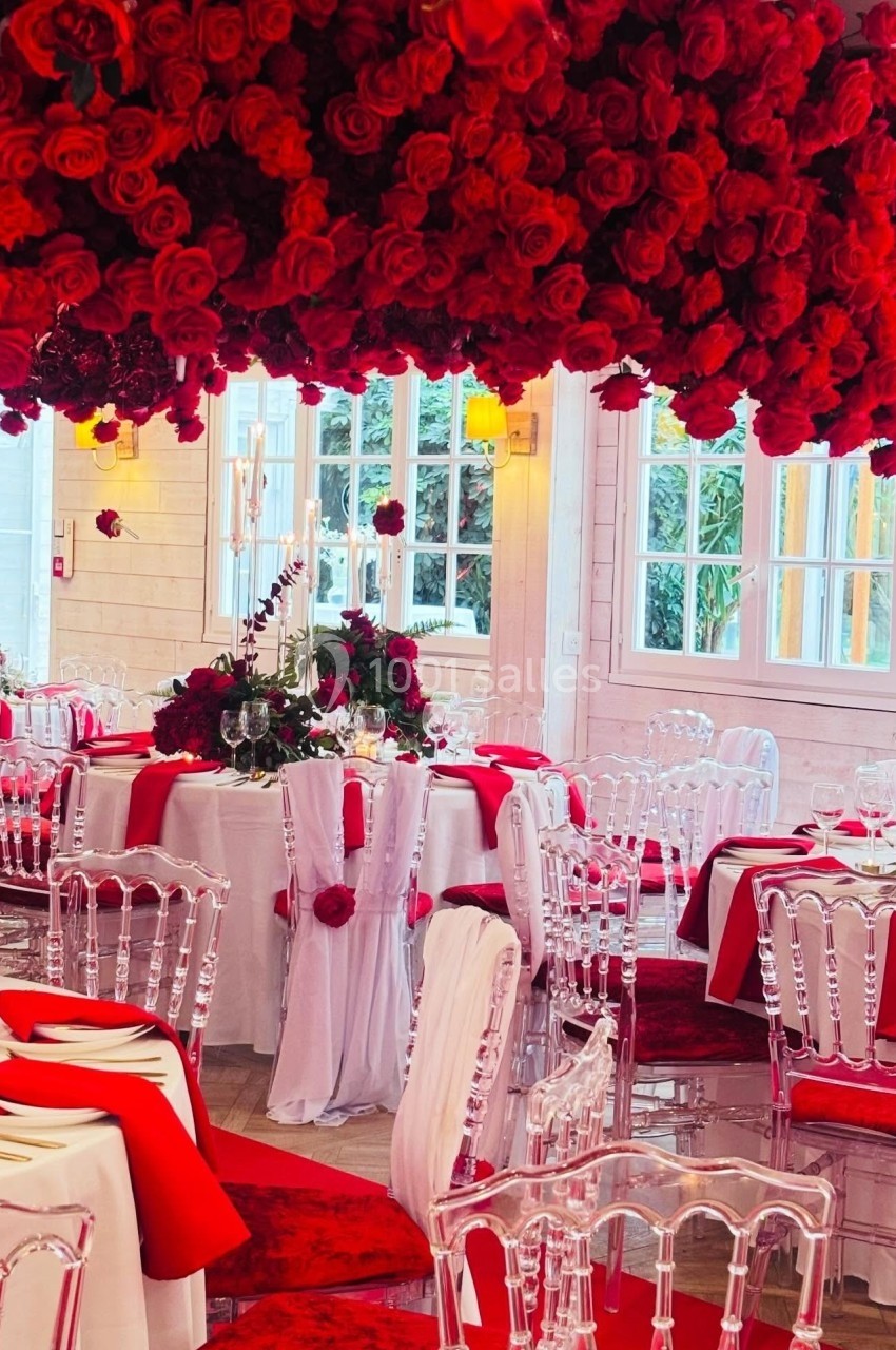 Salle de réception décorée de roses rouges, tables dressées avec nappes blanches et chaises transparentes.