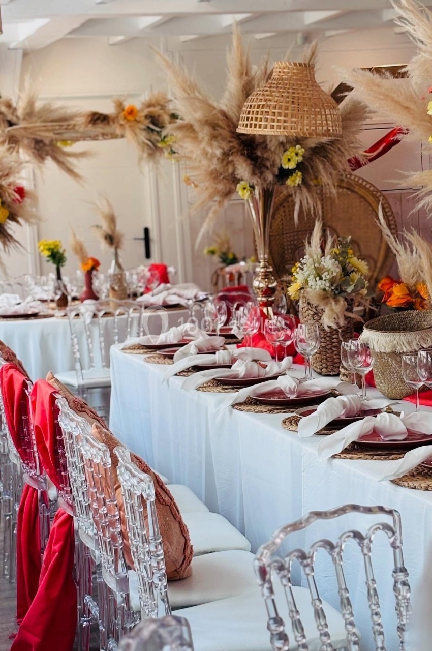 Table décorée pour un événement, avec nappes blanches, chaises transparentes, fleurs séchées et accents rouges.